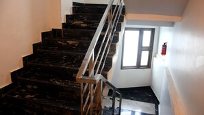 Una scala con gradini in marmo nero e corrimano in metallo, che porta a un pianerottolo con una finestra e un estintore fissato al muro.