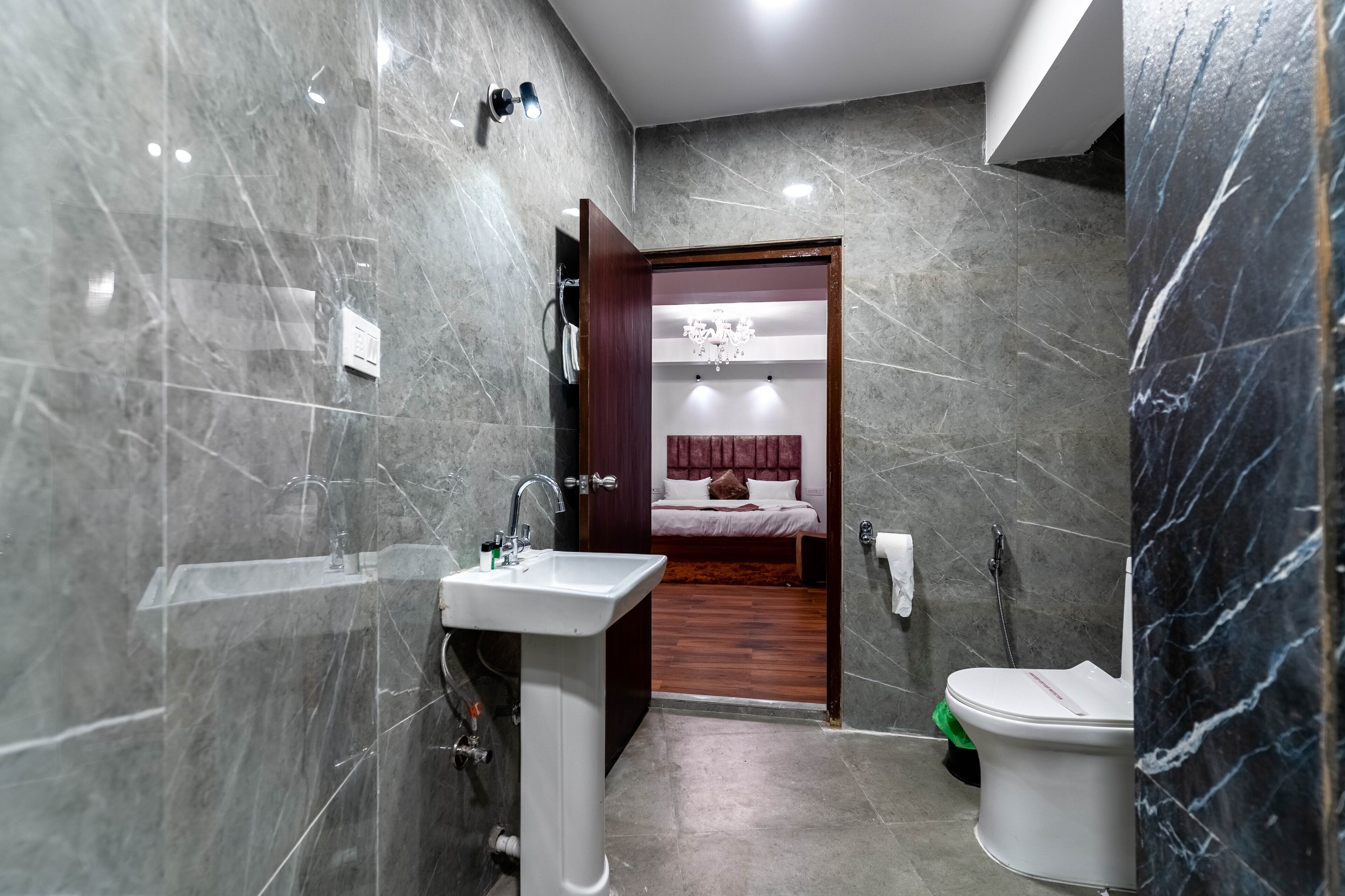 A modern bathroom with a marble wall, a pedestal sink, a toilet, and a wooden door leading to a bedroom with a bed and a chandelier.