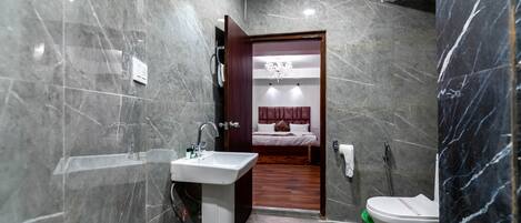 A modern bathroom with a marble wall, a pedestal sink, a toilet, and a wooden door leading to a bedroom with a bed and a chandelier.