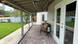 Ein überdachter Patio-Bereich mit Backsteinboden, einer Sitzecke mit Sofa und Stühlen sowie einer Schiebetür zum Garten.