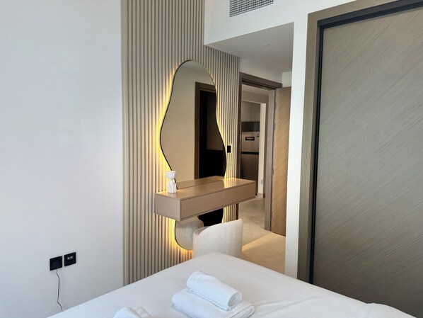 A modern hotel room with a large bed, a bedside table, a mirror, and a view of the kitchen area.