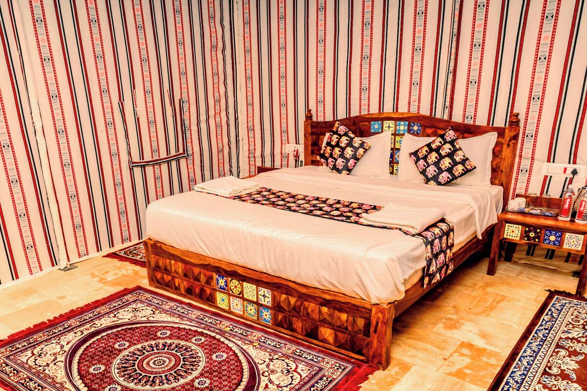 A neatly made bed with a patterned bedspread, a wooden headboard, and decorative pillows. There is a nightstand with a lamp and a water bottle. The room has a patterned carpet and a large, ornate rug.