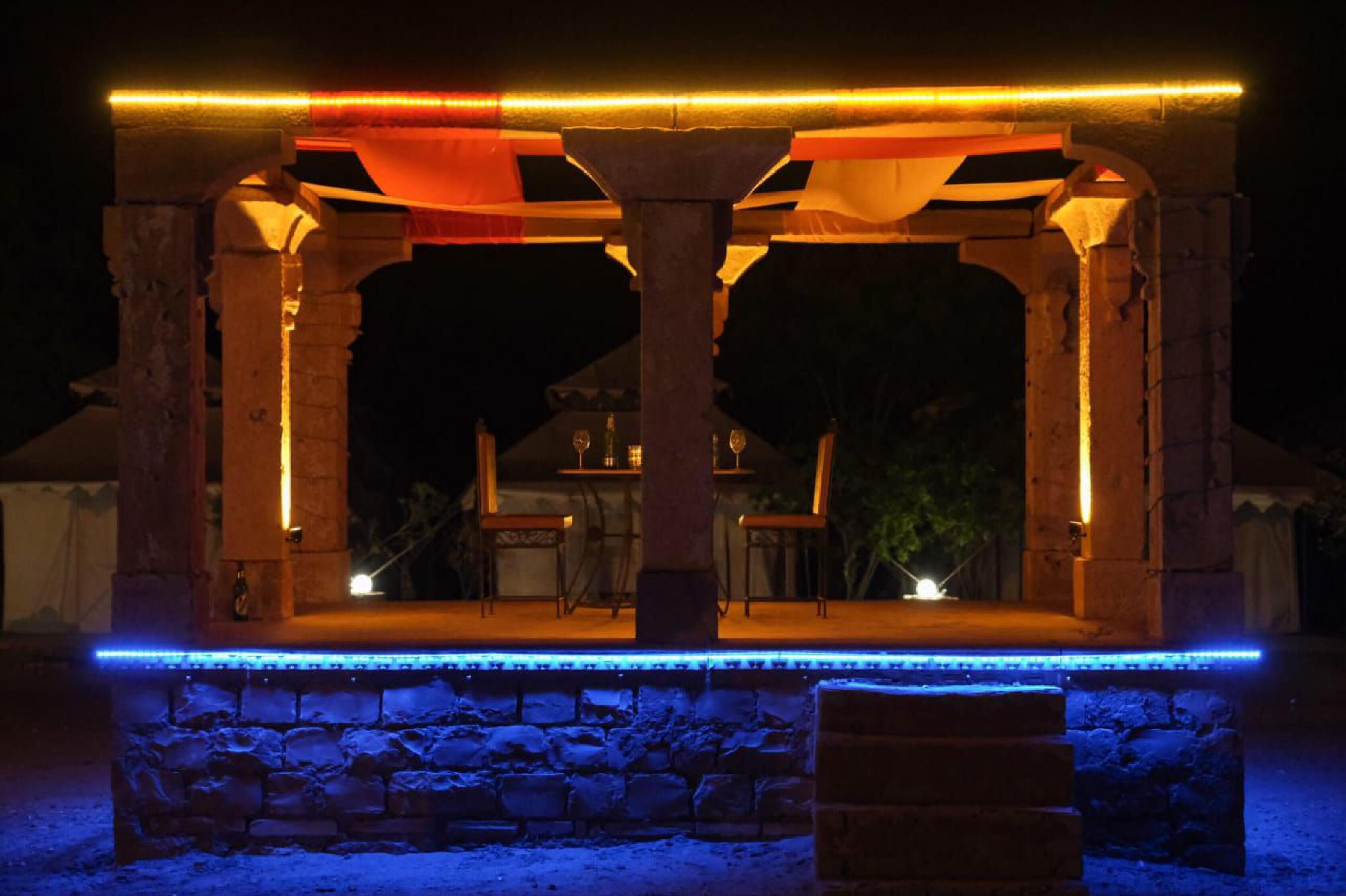 An outdoor seating area with stone columns and a wooden table, illuminated by warm lighting.