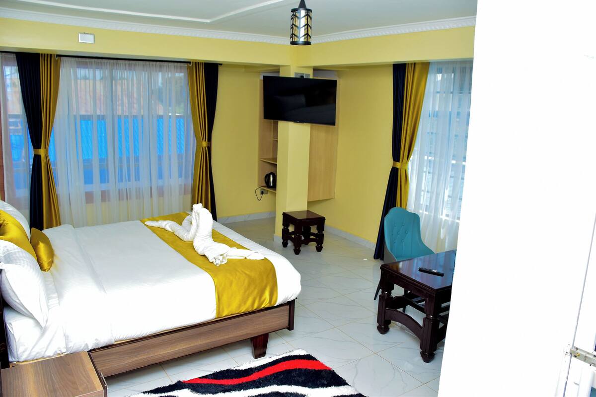 A hotel room with a large bed, a TV mounted on the wall, a small table, a chair, and a view of the outdoors through the windows.