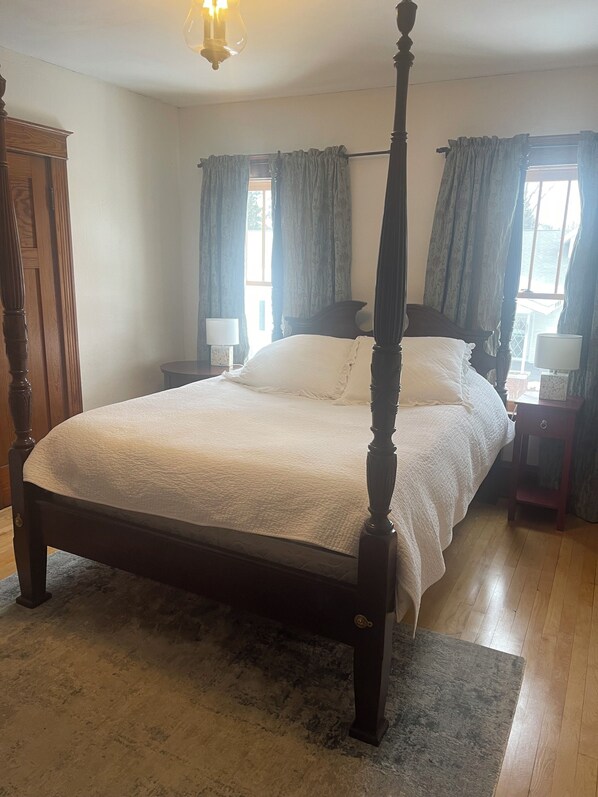 A four-poster bed with a white bedspread and pillows, flanked by two nightstands with lamps, in a room with wooden flooring and a view of the outdoors through a window with curtains. - Vintage Guest House (Burlington)