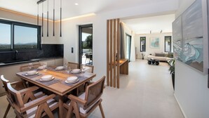 A modern dining area with a wooden table set for six, a kitchen with a large window, and a living area with a sofa and artwork.