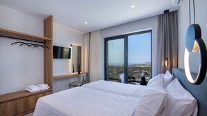 A hotel room with a large bed, a TV mounted on the wall, a wooden bench with towels, and a large window offering a view.