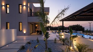 A modern building with multiple illuminated windows, a pool, and a patio area with plants and seating.