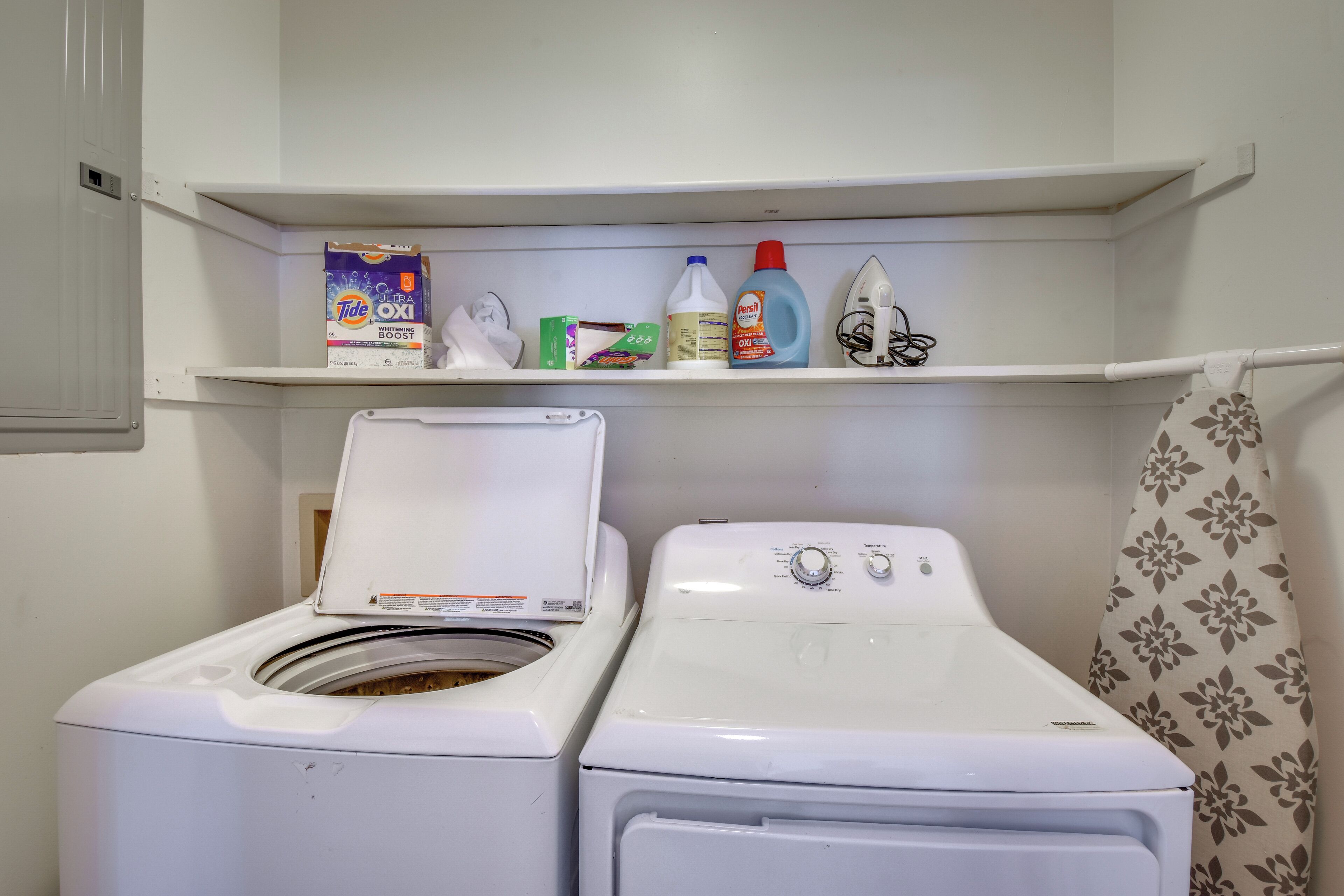 A laundry area with two front-loading washing machines, a shelf with laundry supplies, and a shower curtain.