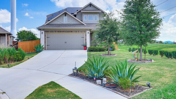 A residential house with a garage, a driveway, and a well-maintained lawn with various plants. - Large Fenced Yard: Family-friendly Kyle Home (Kyle)