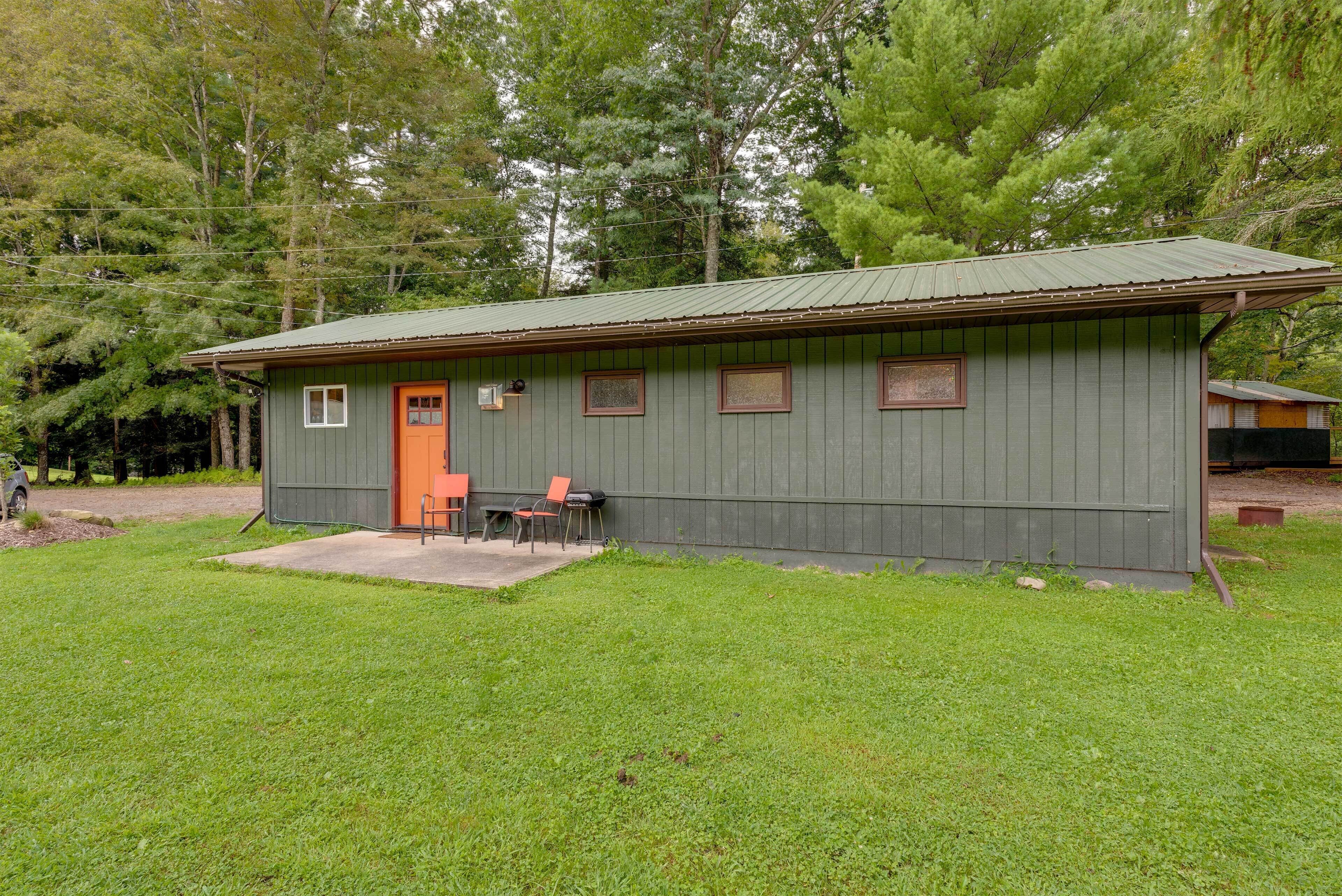 A small, single-story cabin with a green exterior and an orange door, surrounded by a grassy yard and trees.