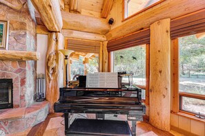 A log cabin interior with a piano, stone fireplace, and large windows offering a view of the outdoors. - Scenic Payson Cabin! Fireplace + Wraparound Patio (Payson)