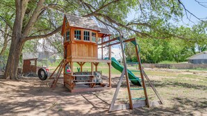 A wooden playhouse with a slide and swings, surrounded by trees and open space. - Family-friendly Albuquerque Home Near Rio Grande! (Albuquerque)