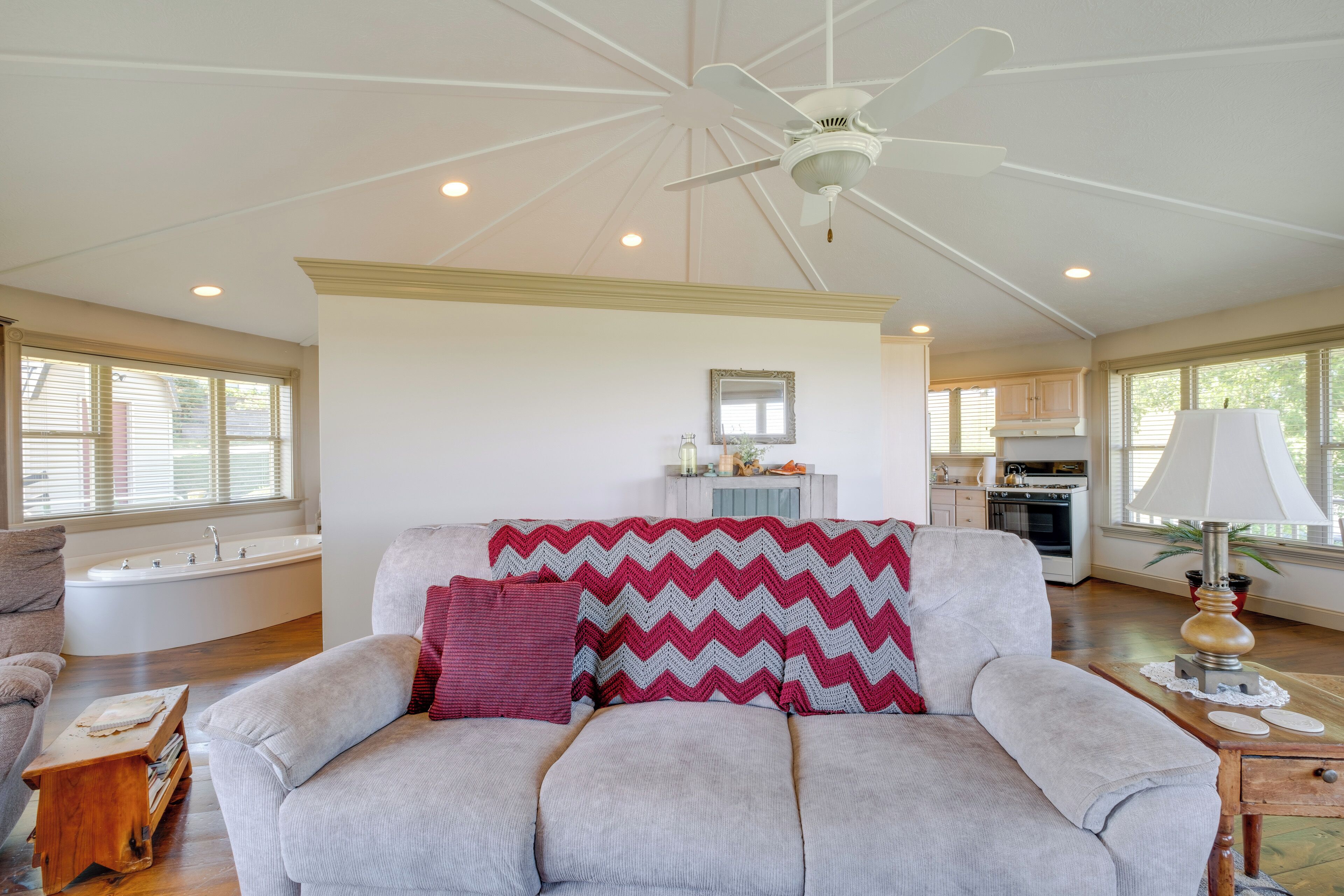A spacious living room with a large grey sofa, a red and grey chevron throw blanket, and a wooden side table with a lamp.