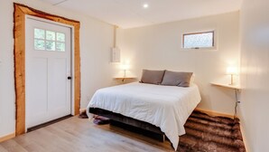 Une chambre avec une porte en bois, un lit avec une literie blanche, deux lampes de chevet, une petite fenêtre et un radiateur fixé au mur.