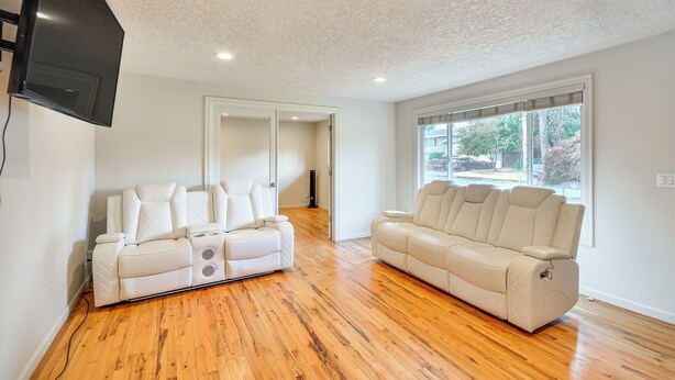 A spacious living room with a large window, two white leather sofas, and a flat-screen TV mounted on the wall. - Milwaukie Home w/ Outdoor Dining Space (Milwaukie)
