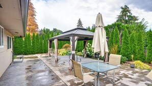 A covered patio with a table and chairs, surrounded by lush greenery and tall trees. - Milwaukie Home w/ Outdoor Dining Space (Milwaukie)