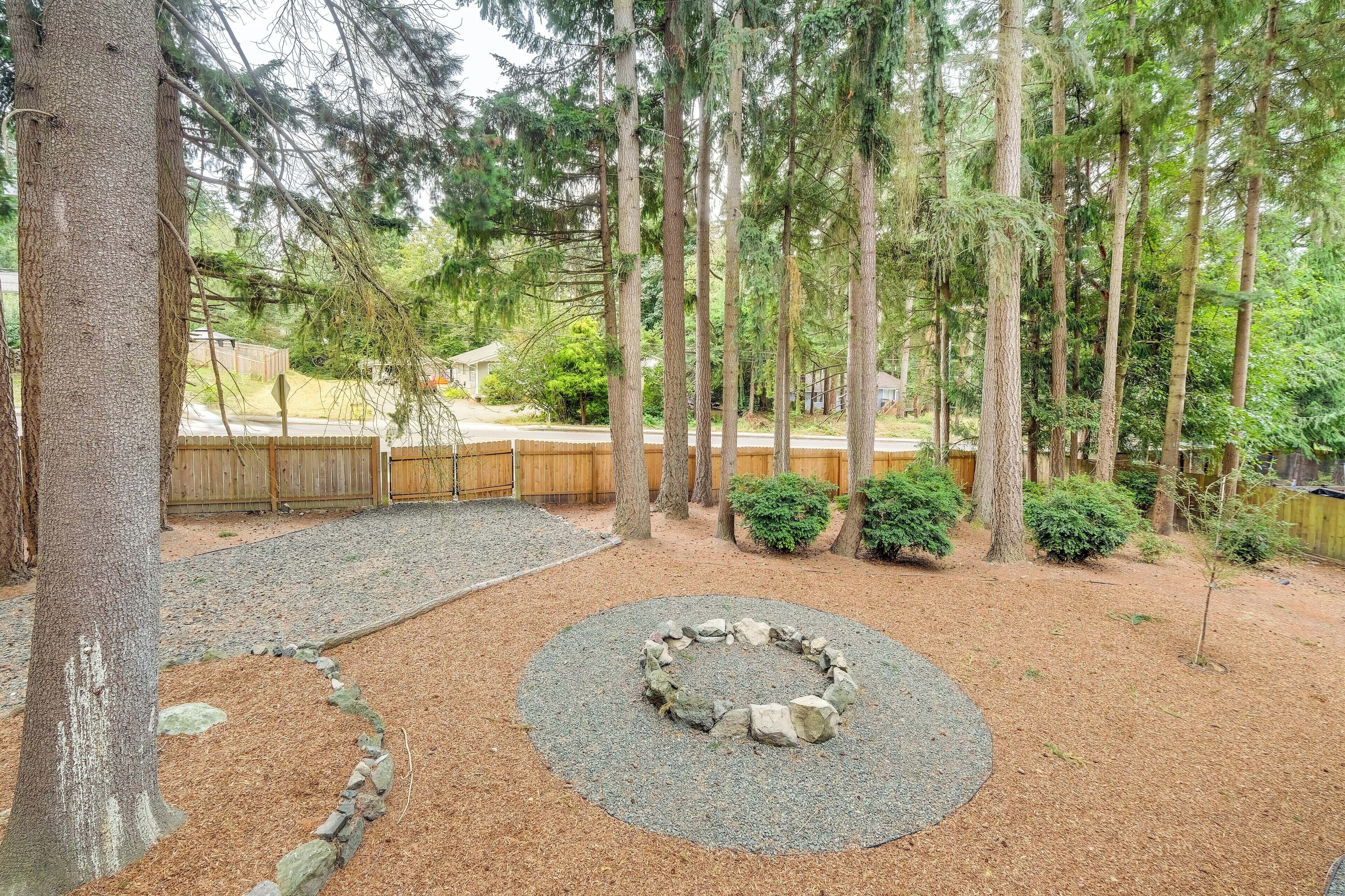 A backyard with a stone fire pit, surrounded by mulch and gravel, with tall trees and a wooden fence in the background.