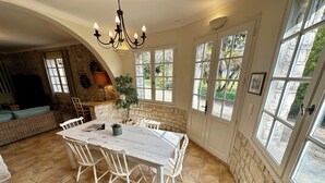 A dining area with a white table and chairs, a chandelier, and large windows with views of greenery.