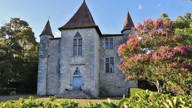 A stone building with a central entrance, two towers, and a garden with pink flowering trees.