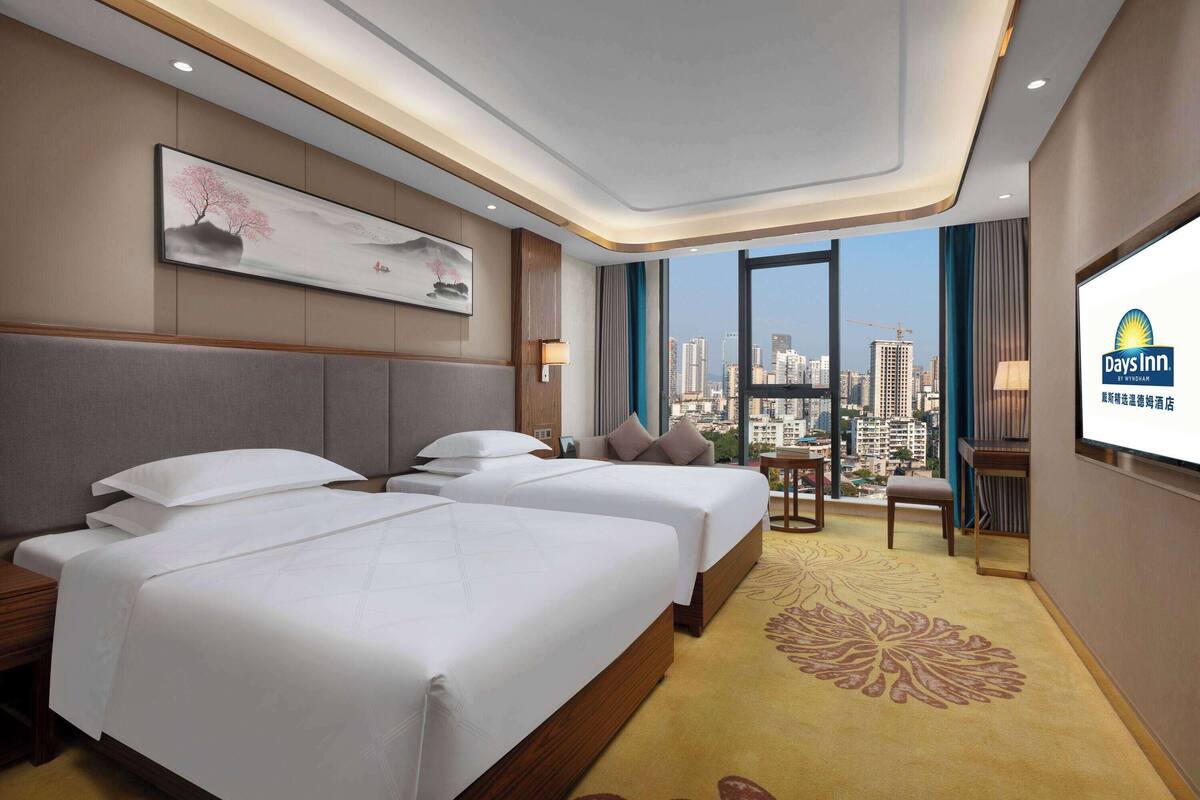 Habitación de hotel con dos camas, un ventanal amplio con vista a la ciudad, televisión y una alfombra decorativa.