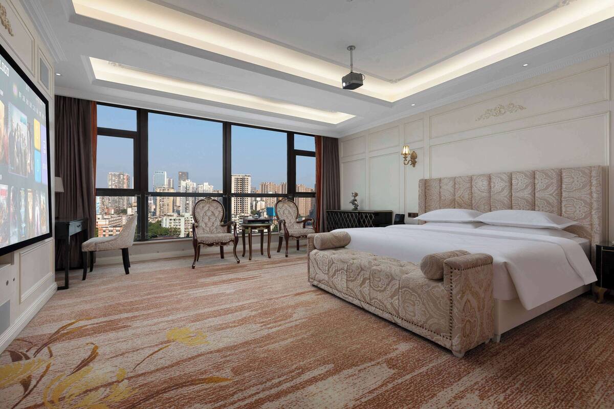 Una habitación de hotel espaciosa con una cama grande, un área de descanso con sillas, un escritorio y un ventanal con vistas a la ciudad.