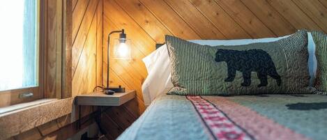 A cozy room with a wooden ceiling, a bed with patterned bedding, a bedside lamp, and a window.
