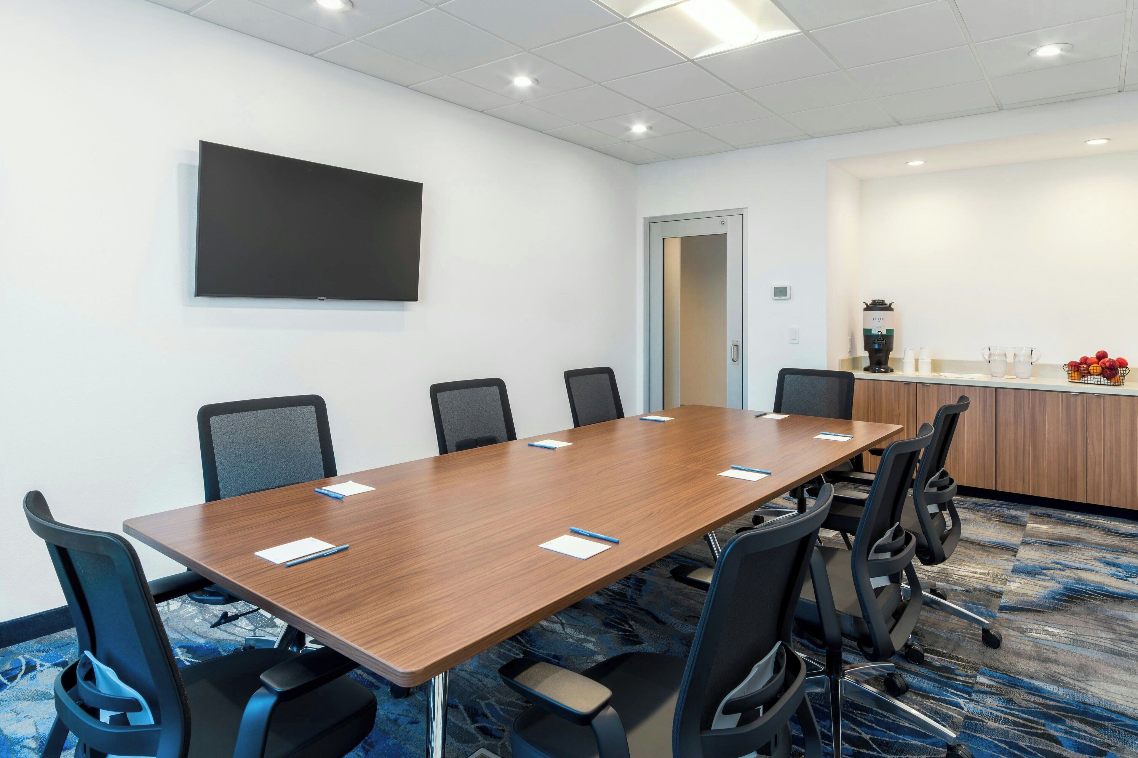 A conference room with a long table, six chairs, a flat-screen TV, a coffee station, and a door.