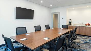 A conference room with a long table, six chairs, a flat-screen TV, a coffee station, and a door.