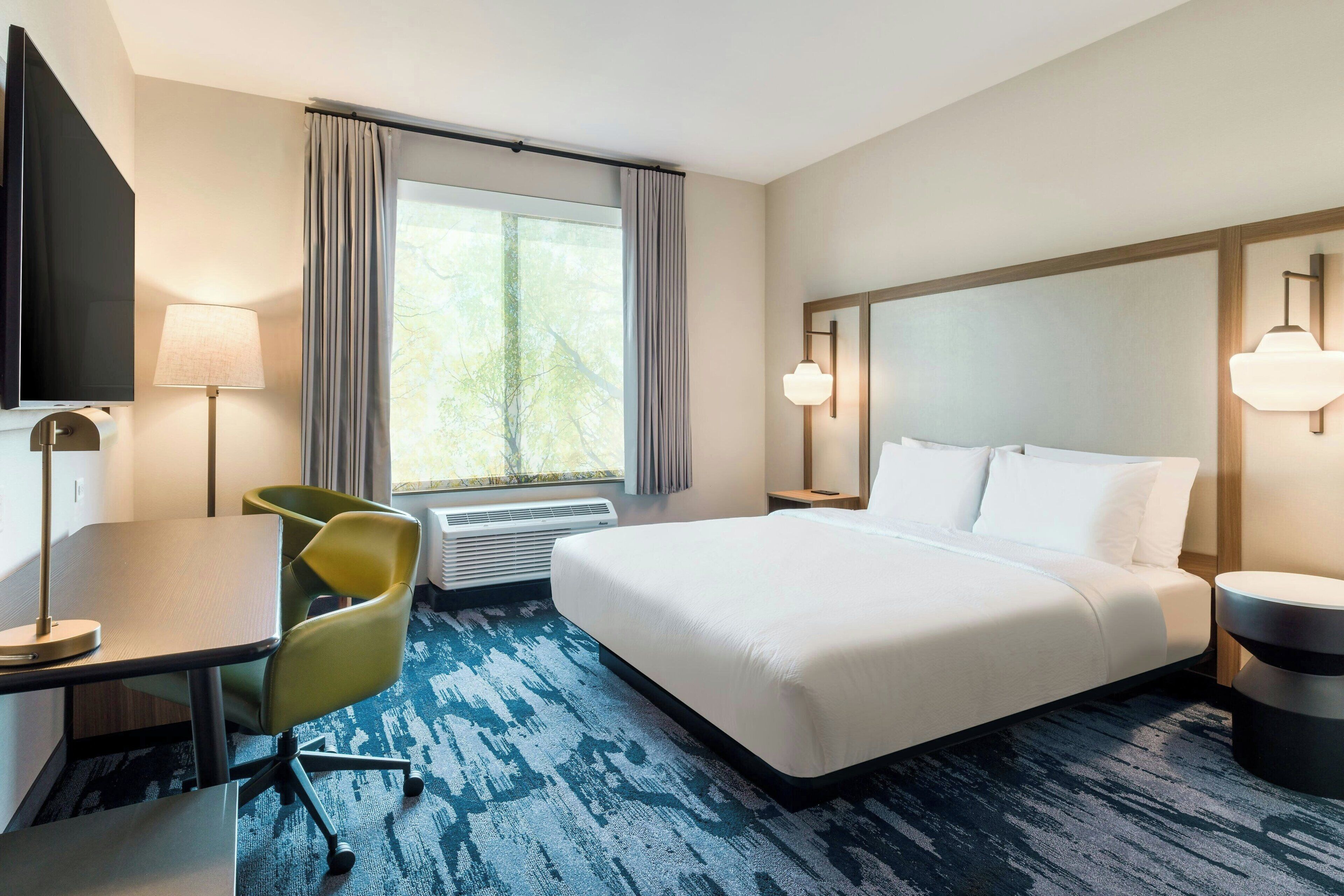 A hotel room with a bed, a desk, a chair, a TV, and a window with curtains.
