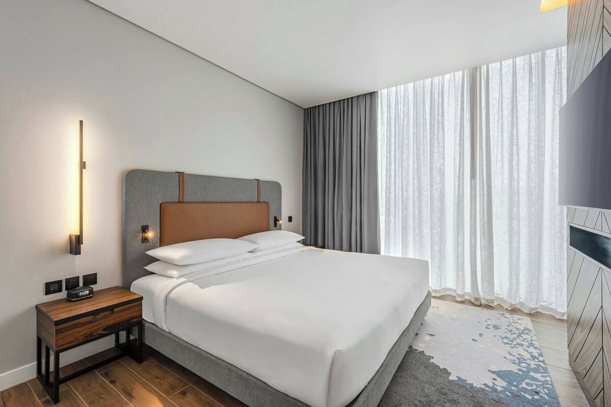 A modern hotel room with a neatly made bed, a bedside table with a lamp, and a large window with curtains.