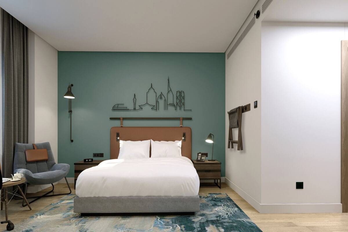 A modern hotel room with a large bed, a cityscape artwork on the wall, a desk with a chair, and bedside lamps.