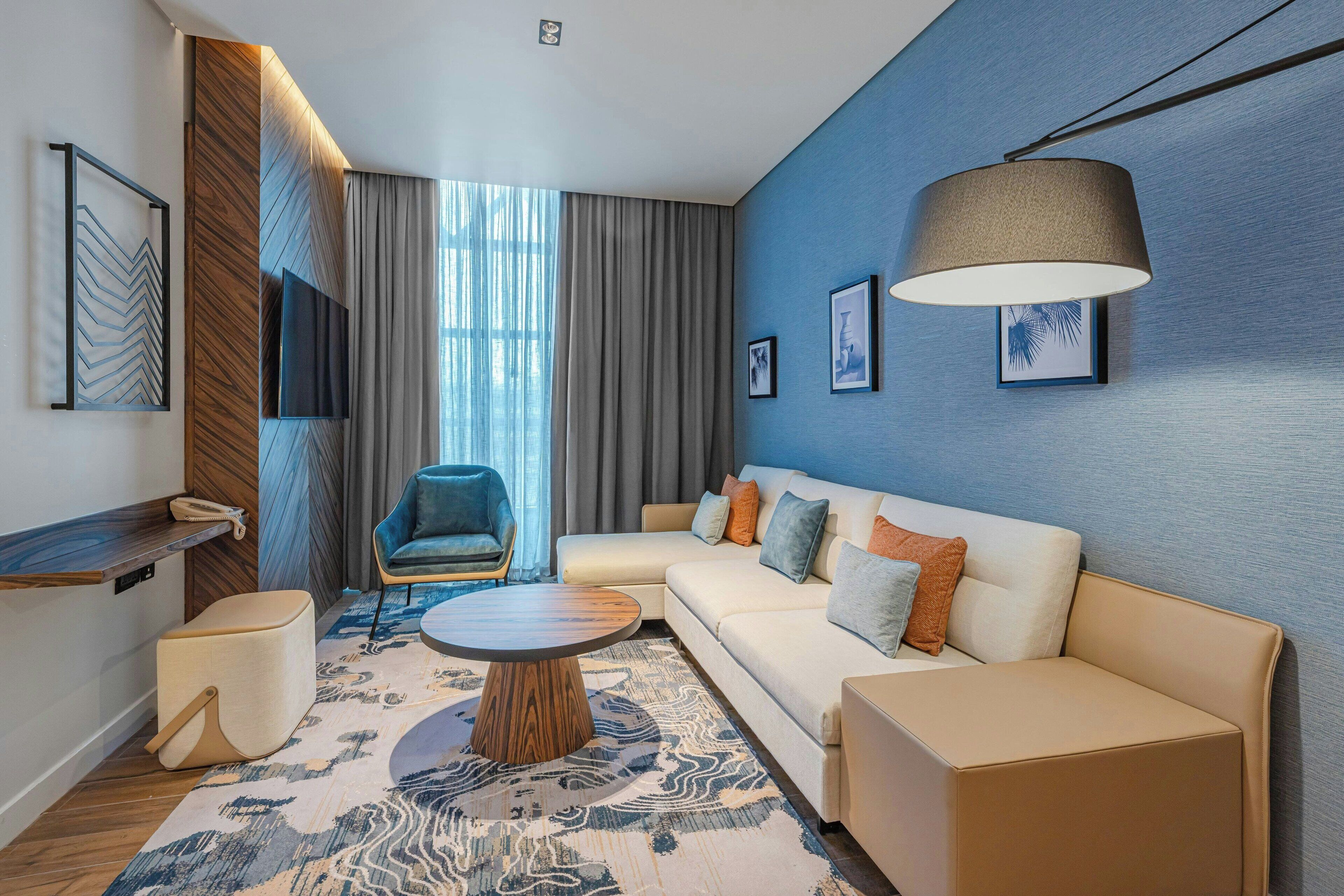 A modern living room with a sectional sofa, a round coffee table, a blue accent wall, and a large window with curtains.