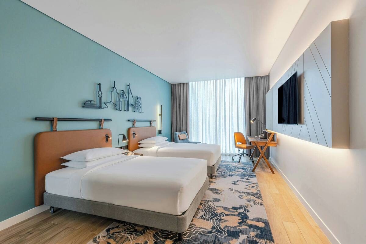 A hotel room with two beds, a desk, a chair, a TV, and a cityscape artwork on the wall.