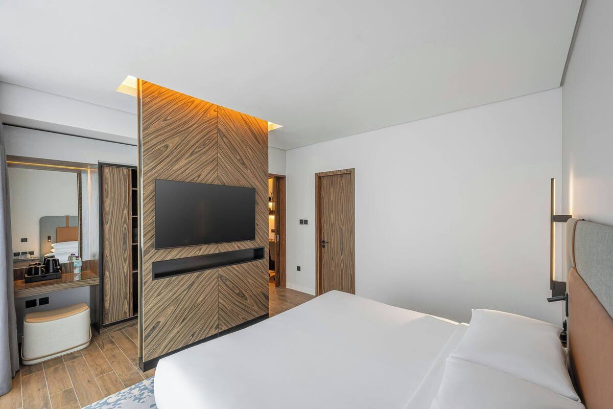 A modern hotel room with a large bed, a flat-screen TV mounted on a wooden panel wall, a bedside table with a lamp, and a mirror.
