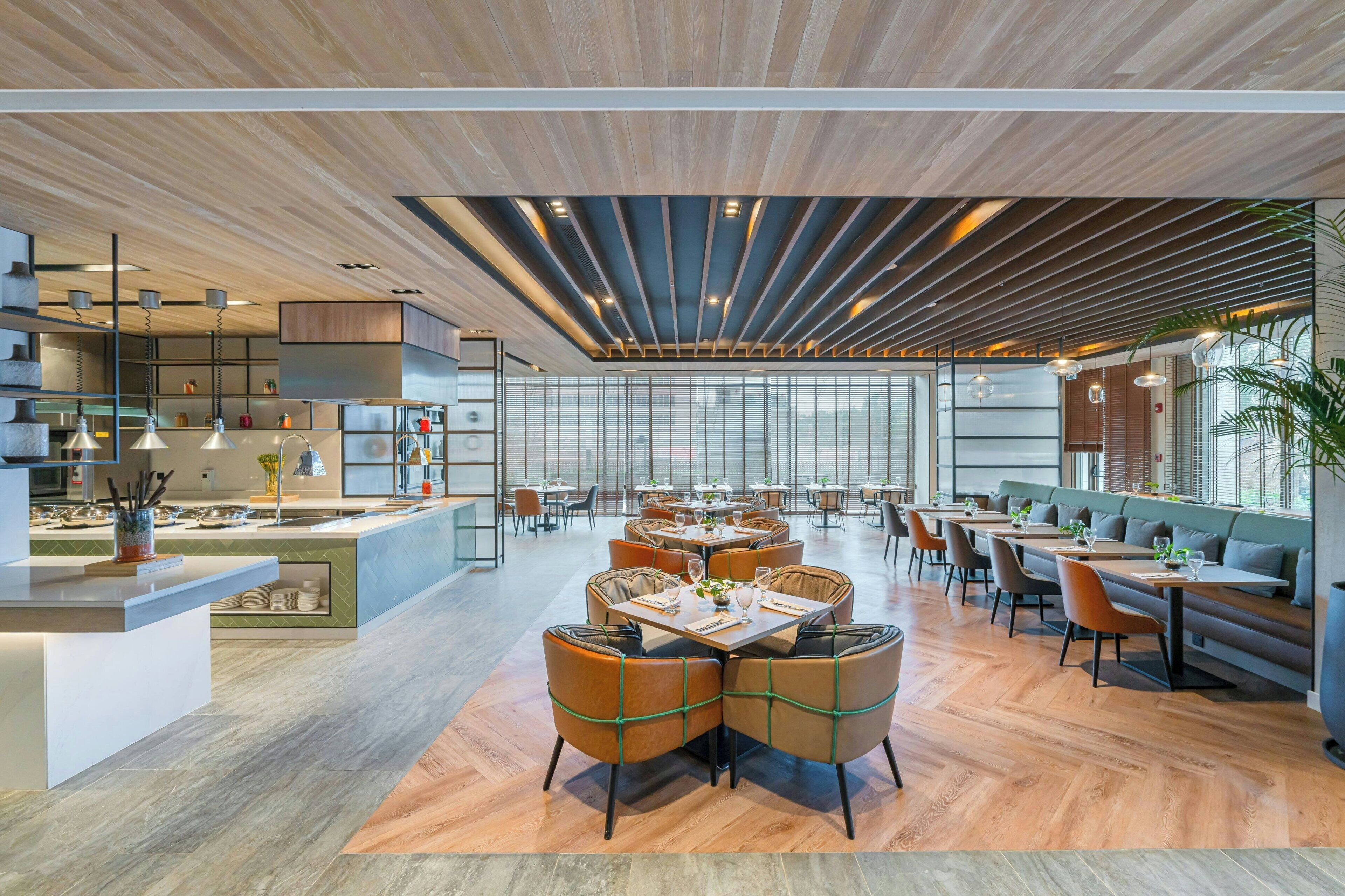 A modern restaurant with wooden flooring, a central dining area with round tables and cushioned chairs, and a bar area with a counter and stools.