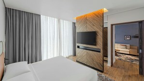 A modern hotel room with a large bed, a flat-screen TV mounted on a wooden panel wall, and a separate seating area with a sofa and coffee table.