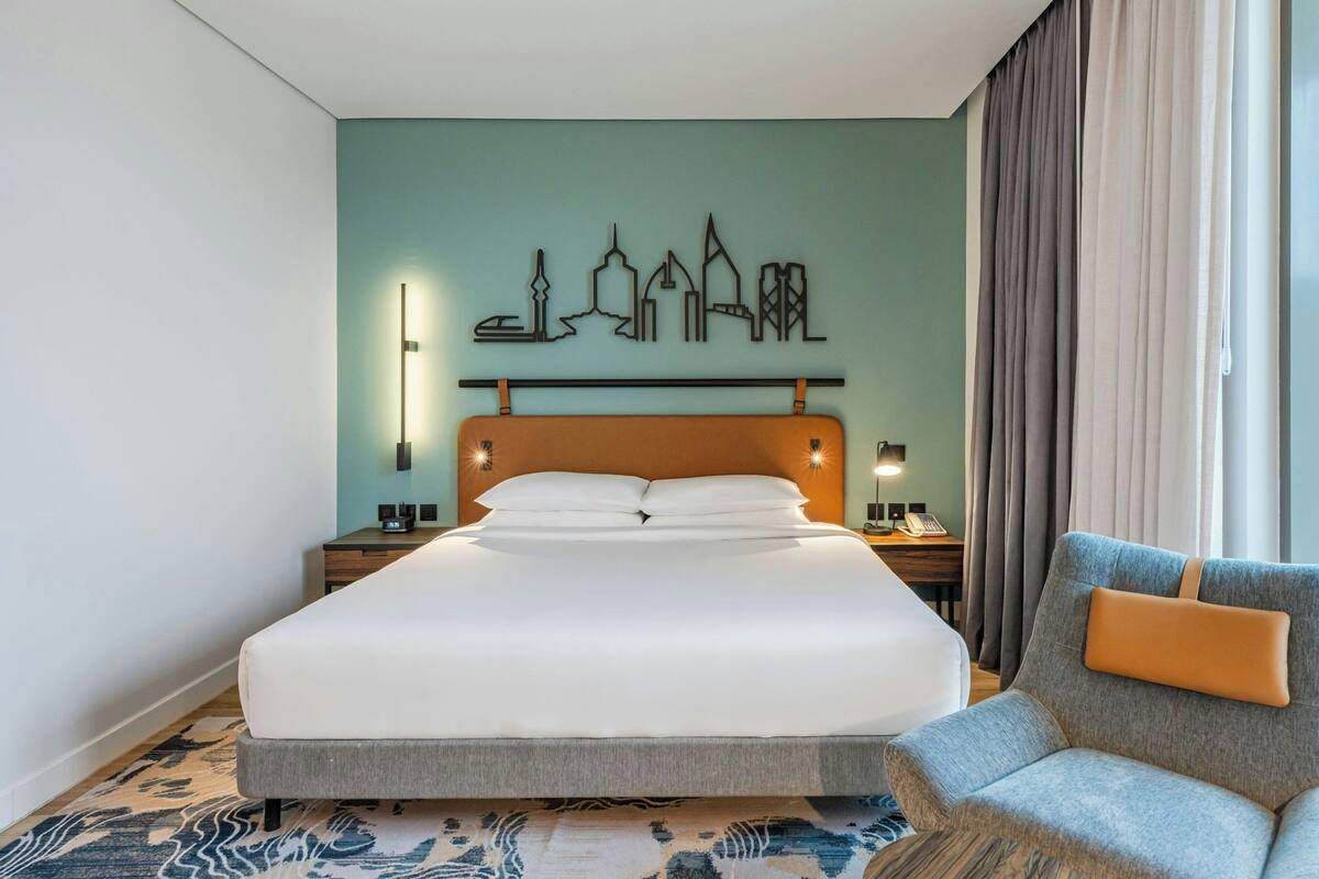 A hotel room with a bed, a chair, a cityscape artwork on the wall, and a nightstand with a lamp.