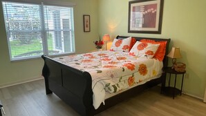 Een slaapkamer met een bed, een raam met zonwering, een lijst met een afbeelding aan de muur en een nachtkastje met een lamp.
