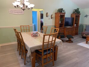 A dining area with a table set for six, surrounded by wooden chairs. A chandelier hangs above the table, and a sign on the wall reads "COME GATHER AT OUR TABLE faith, family, friends."
