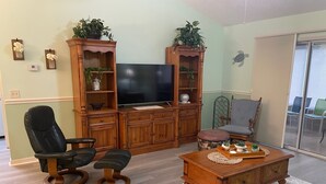 Een woonkamer met een televisie, een houten tv-meubel, twee leren fauteuils, een salontafel en een schuifpui naar een balkon.