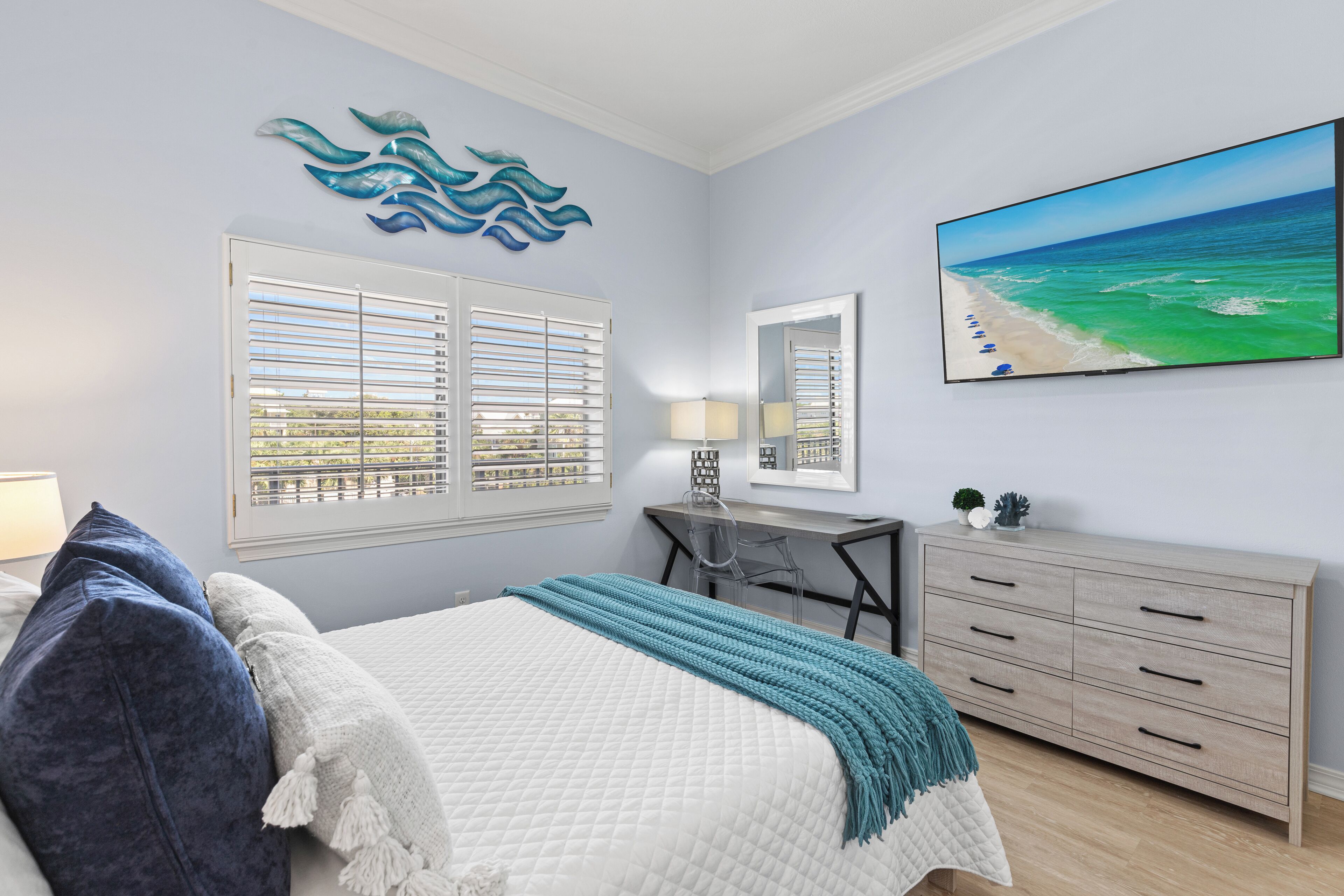 Ein Schlafzimmer mit Bett, Schreibtisch, Kommode, einem Fenster mit Jalousien, einem an der Wand montierten Fernseher und einem Gemälde mit Strandblick.