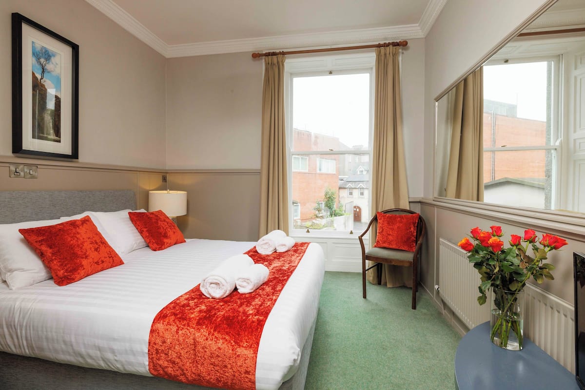 Ein ordentlich bezogenes Bett mit weißer und roter Bettwäsche, ein Blumenstrauß in einer Vase auf einem Tisch und ein Blick auf Gebäude durch das Fenster.