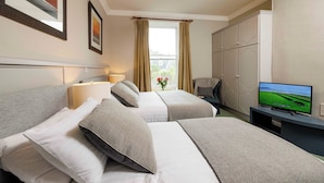 A hotel room with two beds, a TV, a window with curtains, and a view of greenery outside.