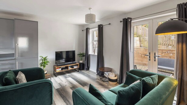 Un salon moderne doté d’un téléviseur à écran plat, de canapés verts, d’une table basse en bois et d’une porte-fenêtre coulissante donnant sur un espace extérieur.