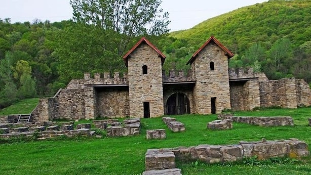 Un edificio de piedra con dos torres y una entrada central, rodeado de campos verdes y árboles.