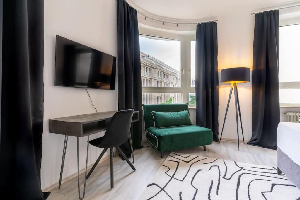 A hotel room with a desk, chair, green sofa, TV, lamp, and a view of buildings.