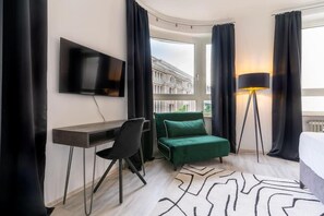 A hotel room with a desk, chair, green sofa, TV, lamp, and a view of buildings.