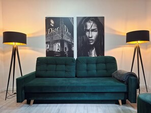 A green sofa with a cushion, two black floor lamps, and a Dior Dior poster on the wall.