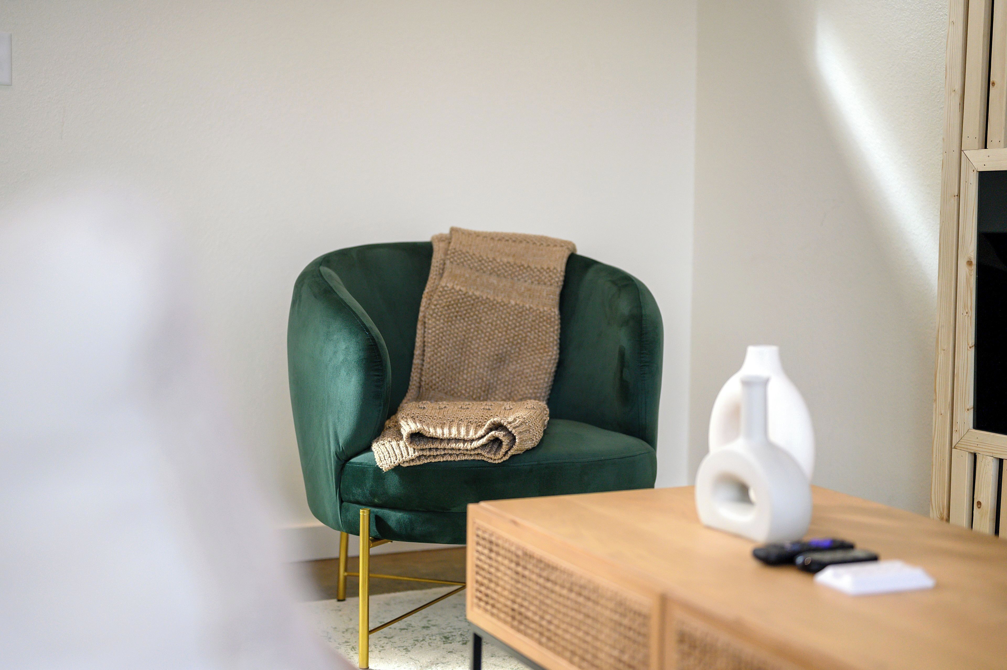 Un fauteuil en velours vert agrémenté d’un plaid tricoté, un vase blanc et une table d’appoint en bois avec des télécommandes.